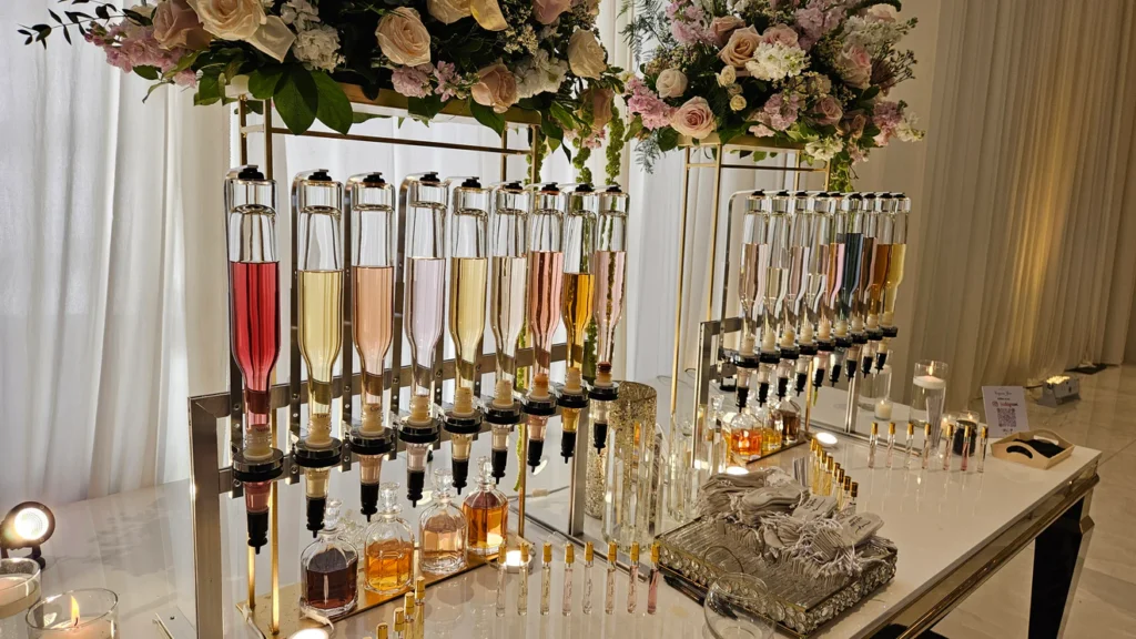 Floral perfume bar at an event Perfume bar with glass dispensers under a floral arrangement of pink and cream roses on a white event table