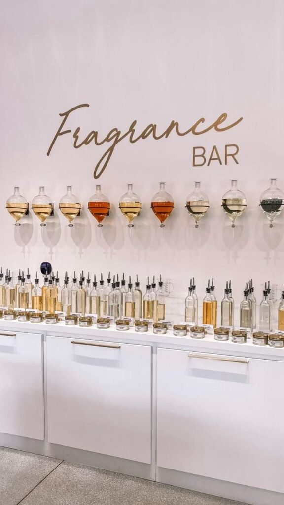 Pink wall with a script Fragrance Bar sign above a row of perfume dispenser bottles on white cabinets, a soft floral setting for sweet fruity scents